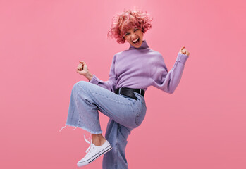 Wall Mural - Charming pink-haired woman in jeans and purple sweater dances on isolated. Curly young active emotional girl move on pink background.