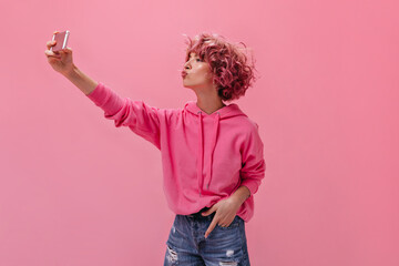 Wall Mural - Pretty woman in fuchsia color hoodie and denim shorts blows kiss and makes selfie on isolated. Portrait of cool young girl holding phone on pink background.