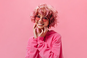 Wall Mural - Cheerful happy woman in pink sunglasses poses on isolated background. Portrait of joyful girl in hoodie on pink backdrop.