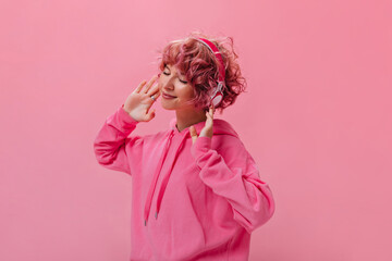 Wall Mural - Happy young curly girl in pink hoodie dance and listens to music in headphones on isolated. Short-haired girl smiles sincerely on pink background.