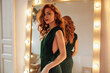 © Look! - Cheerful woman in dark green outfit poses near mirror. Lovely lady with ginger wavy hair in stylish dress looking into camera..