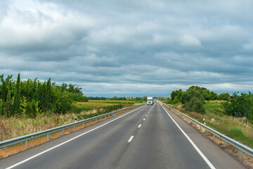 Wall Mural - Landscape of a road lined with trees, cultivated fields and a cloudy sky, with a truck driving straight ahead.