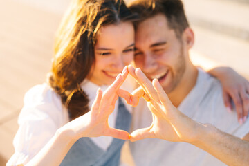 Close-up of happiness young couple making heart with hands resting in city park in summer sunny day. Relationships happy boyfriend and girlfriend enjoying spending time together outdoors.