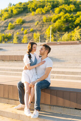 Full length vertical portrait of happy young couple resting in city park sitting on bench in summer sunny day. Relationships between cheerful boyfriend and girlfriend enjoying time together outdoors.