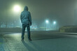 A mysterious hooded figure with back to camera, standing on a road in a light industrial urban area. On a moody, foogy, winters night