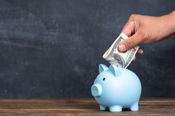 A man's hand puts a 100 dollar bill into a blue piggy bank.