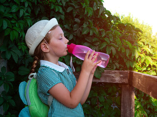 Cute school little girl drinks water from reusable pink bottle outdoor. Child in hat enjoys fresh cold water on green summer street. Body rehydration hot weather, human organism liquid assets keeping