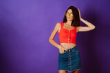 Fototapeta Panele - Beautiful young woman in a red top, skirt and yellow glasses posing on a purple background. Beauty and positive concept.
