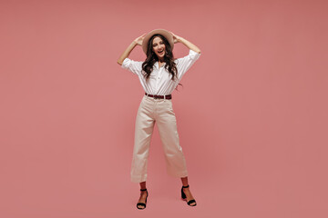 Wall Mural - Photo of fashionable lady with curly hair in white shirt, light pants and black heels holding her wide-brimmed hat and smiling..