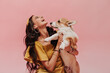 Positive woman with long hair in earrings and bandana in trendy yellow outfit laughing and holding cute dog on pink backdrop..
