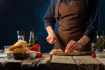 Wall Mural - The process of preparing the classic Caesar salad. The cook cuts the chicken fillet into pieces. Many bright ingredients are on the left side of the photo. Rough wooden table. Country style.