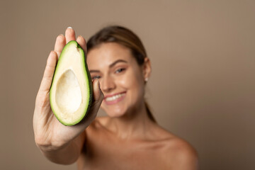 Wall Mural - Happy smiling young woman holding avocado half on a beige background. Natural beauty concept