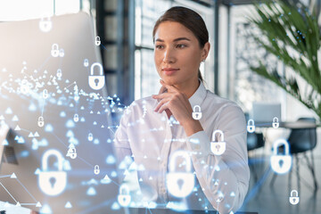 Wall Mural - Attractive businesswoman in white shirt at workplace working with laptop to defend customer cyber security. Concept of clients information protection. Padlock hologram over office background.