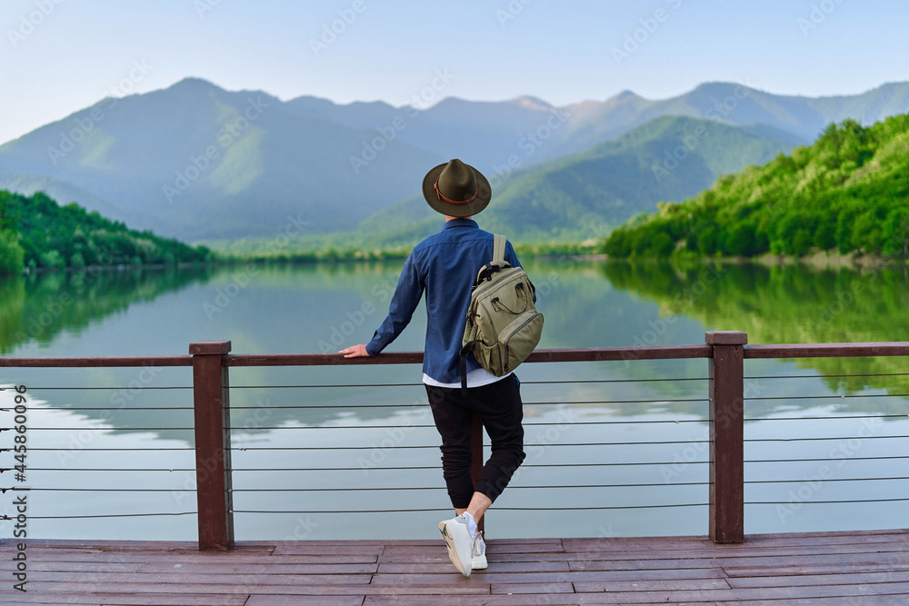 Hipster traveler backpacker standing alone on edge of pier and staring at lake and mountains. Enjoying beautiful freedom moment life and serene quiet peaceful atmosphere in nature. Back view - obrazy, fototapety, plakaty 