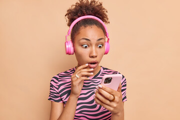 Wall Mural - Surprised curly haired ethnic dark skinned woman stares at smartphone display updates playlist wears stereo wireless headphones striped t shirt isolated over beige background reacts on shocking news