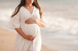 Smiling pregnant woman wear white dress hol tummy walk at beach over sea nature background outdoors. Summer vacation season. Motherhood. Maternity.