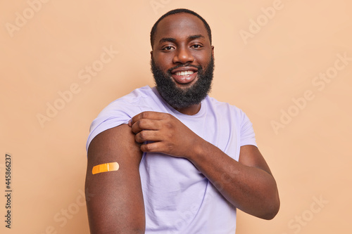Happy dark skinned bearded man shows arm with adhesive plaster got coronavirus vaccine feels safe smiles pleasantly isolated over brown background. Vaccination campaign and immunization concept