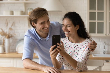 Wall Mural - Laughing millennial spouses embrace at home kitchen enjoy watching cute funny video clips photos pictures at social network together. Happy joyful family couple looking at smartphone screen having fun