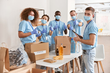 Wall Mural - Group of diverse people wearing blue uniform, protective masks and gloves showing thumbs up while sorting donated food items, volunteering in community together