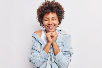 Wall Mural - Pretty curly haired ethnic woman closes eyes smiles broadly keeps hands under chin has gentle expression wears denim jacket isolated over white background feels heartwarming stands romantic.