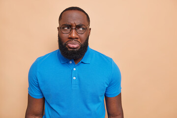 Wall Mural - Unhappy discontent dark skinned man has sulking expression wants to cry because of problems purses lips looks away wears big spectacles basic casual blue t shirt isolated over brown background