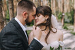 © shchus - Wedding portrait of newlyweds close up. A bearded, stylish groom in a suit and a beautiful brunette bride in a white dress with a bouquet in her hands are standing and hugging in nature in the forest.