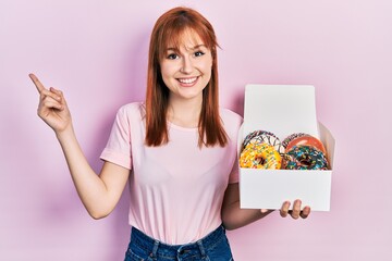 Wall Mural - Redhead young woman holding tasty colorful doughnuts smiling happy pointing with hand and finger to the side