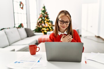 Wall Mural - Young caucasian girl sitting on the table working using laptop by christmas tree smiling with hands on chest with closed eyes and grateful gesture on face. health concept.