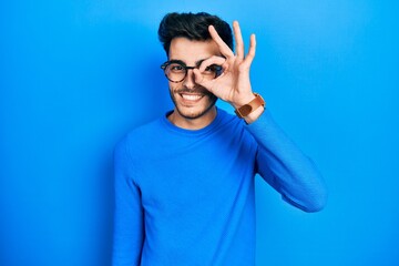 Young hispanic man wearing casual clothes and glasses doing ok gesture with hand smiling, eye looking through fingers with happy face.