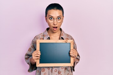 Wall Mural - Beautiful hispanic woman with short hair holding blackboard in shock face, looking skeptical and sarcastic, surprised with open mouth