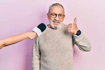 Sticker - Handsome senior man with beard being interviewed by reporter holding microphone smiling happy and positive, thumb up doing excellent and approval sign