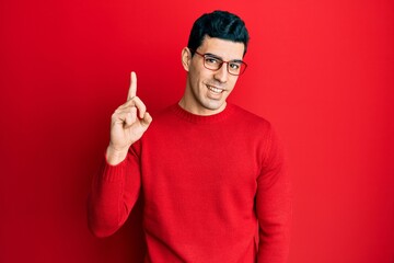 Wall Mural - Handsome hispanic man wearing casual clothes and glasses smiling with an idea or question pointing finger up with happy face, number one