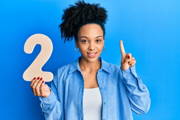 Wall Mural - Young african american girl holding number two smiling with an idea or question pointing finger with happy face, number one