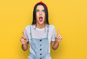 Wall Mural - Young hispanic girl wearing casual clothes amazed and surprised looking up and pointing with fingers and raised arms.