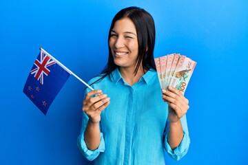 Poster - Young latin woman holding new zealand flag and dollars winking looking at the camera with sexy expression, cheerful and happy face.