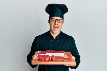 Handsome young man wearing chef uniform holding board with raw meat celebrating crazy and amazed for success with open eyes screaming excited.