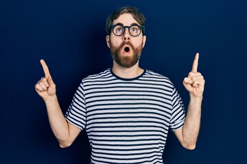 Wall Mural - Caucasian man with beard wearing striped t shirt and glasses amazed and surprised looking up and pointing with fingers and raised arms.