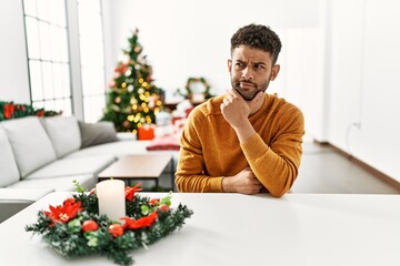 Sticker - Arab young man sitting on the table by christmas tree with hand on chin thinking about question, pensive expression. smiling with thoughtful face. doubt concept.