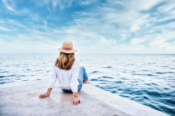 Wall Mural - Rear view shot of a woman wearing straw hat and casual clothes while sitting on seaside and looking at the sea.