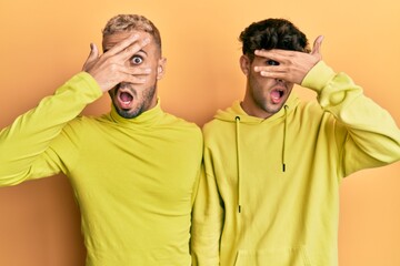 Sticker - Homosexual gay couple standing together wearing yellow clothes peeking in shock covering face and eyes with hand, looking through fingers with embarrassed expression.