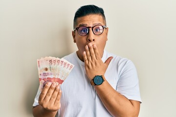 Canvas Print - Young latin man holding 10 colombian pesos banknotes covering mouth with hand, shocked and afraid for mistake. surprised expression