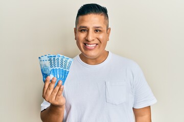 Canvas Print - Young latin man holding 10000 chilean pesos looking positive and happy standing and smiling with a confident smile showing teeth