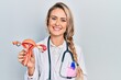 Beautiful young blonde woman holding anatomical model of female genital organ looking positive and happy standing and smiling with a confident smile showing teeth