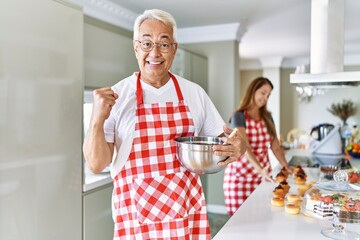 Wall Mural - Middle age hispanic couple wearing apron cooking homemade pastry screaming proud, celebrating victory and success very excited with raised arms