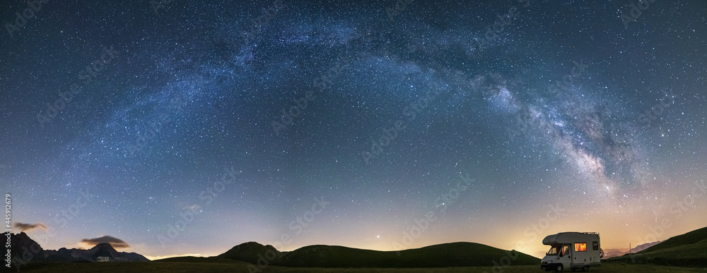 Panoramic night sky over Campo Imperatore highlands, Abruzzo, Italy. The Milky Way galaxy arc and stars over illuminated camper van. Camping freedom in unique hills landscape. - obrazy, fototapety, plakaty 