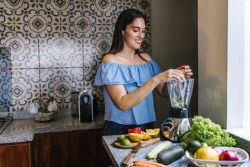 Wall Mural - Latin Woman making green smoothie or Detox juice in kitchen at Home in healthy eating concept in Mexico