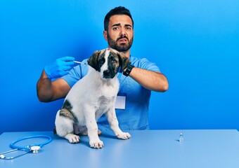 Poster - Handsome hispanic veterinary man with beard putting vaccine to puppy dog clueless and confused expression. doubt concept.