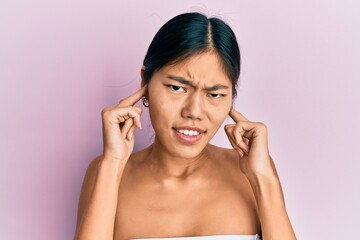 Poster - Young chinese woman standing topless showing skin covering ears with fingers with annoyed expression for the noise of loud music. deaf concept.