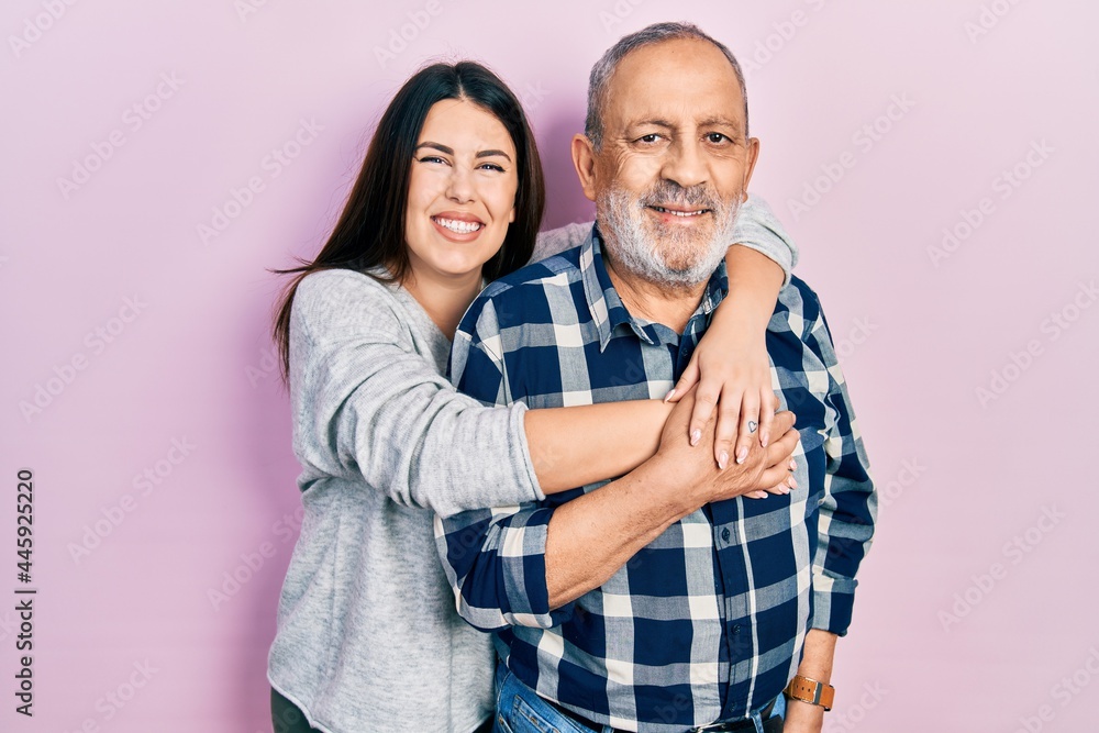 Young brunette woman and senior man standing over pink background. Daughter and father hugging and bonding together as happy family - obrazy, fototapety, plakaty 