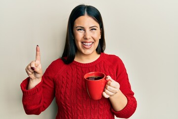 Wall Mural - Beautiful brunette woman drinking a cup of black coffee smiling with an idea or question pointing finger with happy face, number one